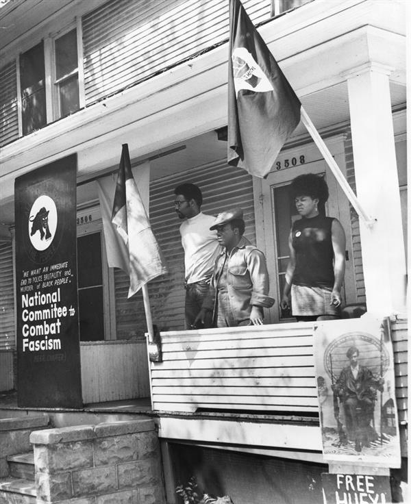 Three black individuals stand on a porch with a sign for National Committee to Combat Fascism